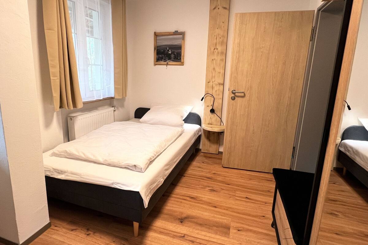 Um quarto pequeno e arrumado, com uma cama, um guarda-roupa de madeira e uma janela com cortinas.