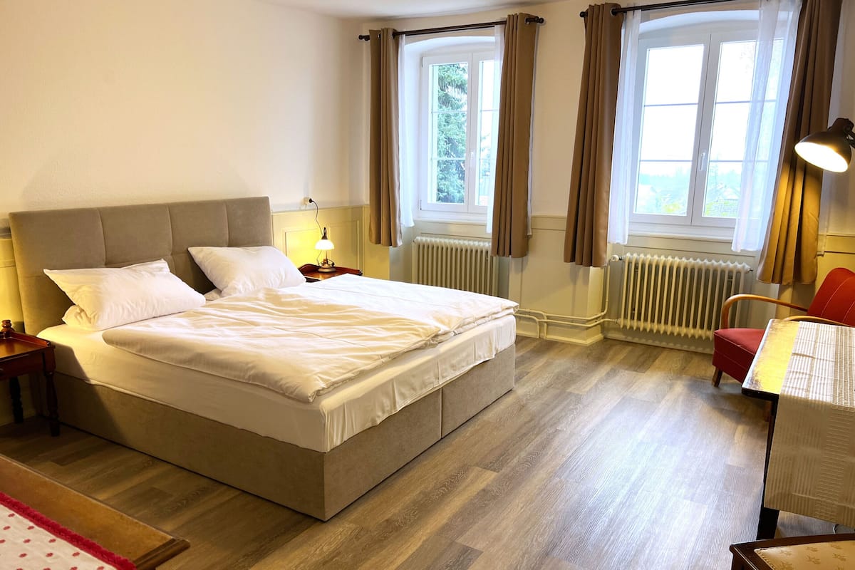 Uma cama bem arrumada com cabeceira, mesa de cabeceira com luminária, uma cadeira e uma janela com cortinas.