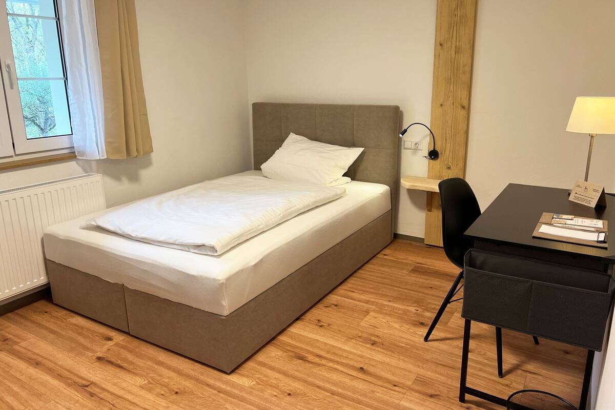 Uma cama de solteiro com cabeceira, uma escrivaninha com cadeira, uma luminária e uma janela com cortinas.