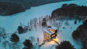 A modern, multi-story house with a flat roof, surrounded by a snowy landscape and dense forest. - Lakeside Loft, Roof Deck + Sauna (Phelps)