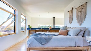 A bedroom with a wooden bed, a view of the outdoors through large windows, a desk with a plant, and macrame wall art. - Lakeside Loft, Roof Deck + Sauna (Phelps)