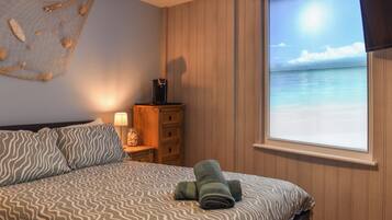 A bedroom with a bed, a nightstand, a dresser, a window with a beach view, and a wall decoration.