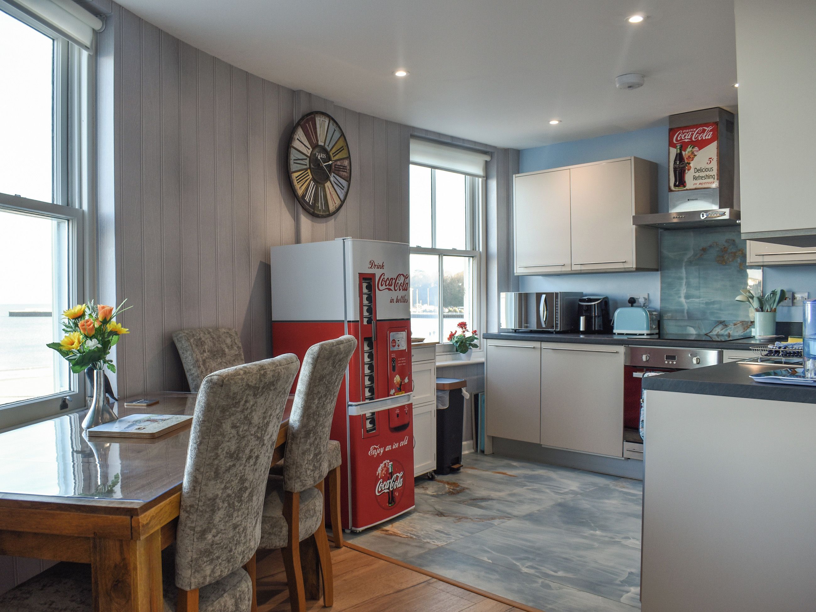 A modern kitchen with a vintage Coca-Cola refrigerator, a dining area with a vase of flowers, and a large wall clock.
