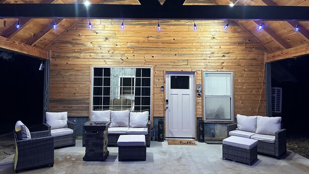 An outdoor covered patio with wooden walls, a white door, and two windows. There are wicker sofas with cushions, a stone fireplace, and a concrete floor.
