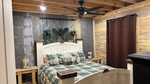 A bedroom with a wooden ceiling, a bed with plaid bedding, a nightstand with a lamp, and a window with a curtain.