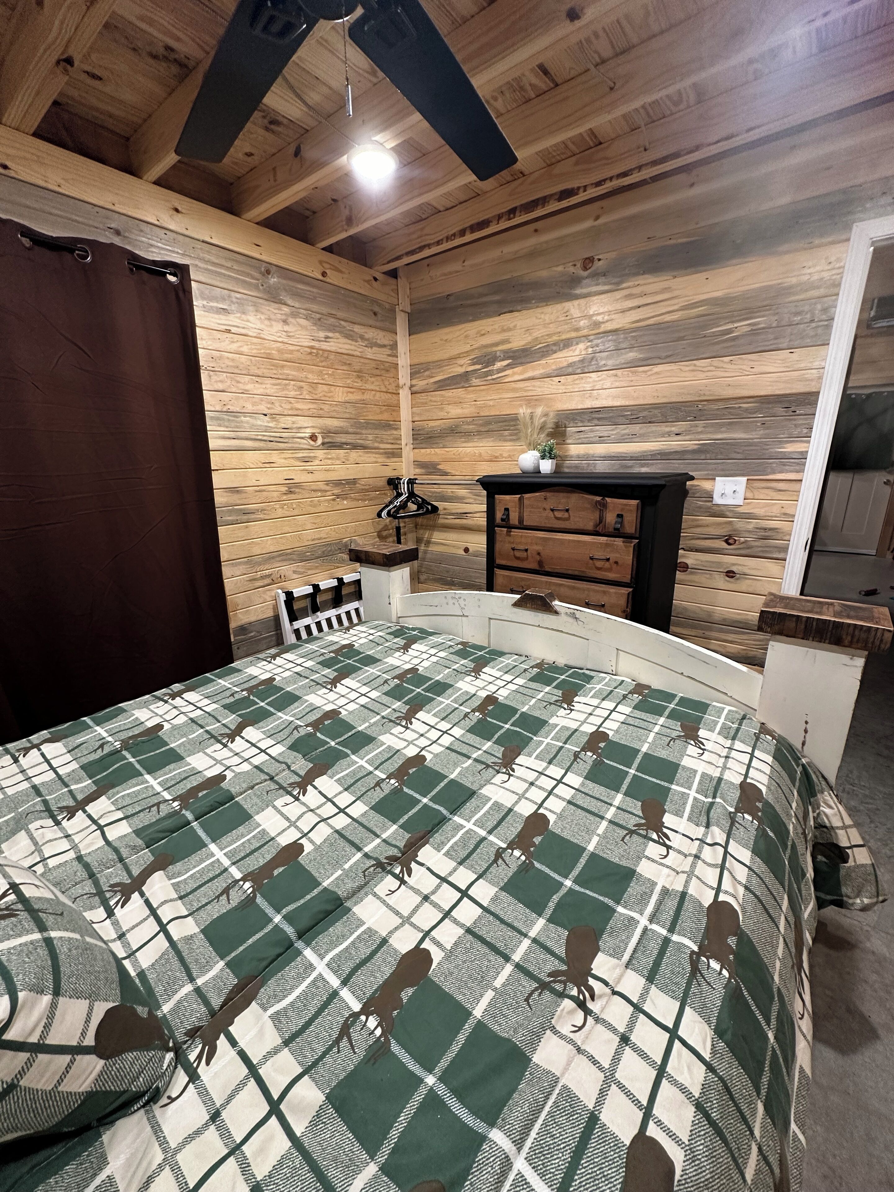 A bedroom with a wooden ceiling, a bed with a checkered bedspread, a dresser, and a small table.