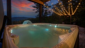 Un jacuzzi bajo una pérgola de madera con luces colgantes, rodeado de árboles y vistas al paisaje.
