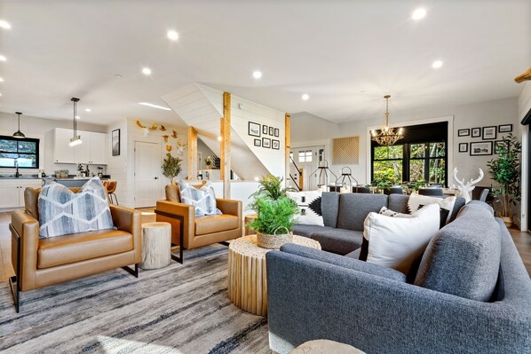 A spacious living area with a sofa, armchairs, and a coffee table. There is a staircase leading to an upper level, and a dining area with a chandelier.