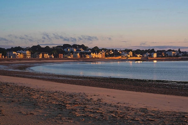 A coastal beach scene with a wide sandy shore, calm water, and a row of buildings with lights on in the background. - Steps from the ocean - minutes from Boston (Winthrop)