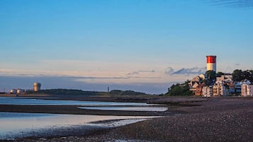 Ett kustlandskap med en strand, en fyr och industriella byggnader i bakgrunden.