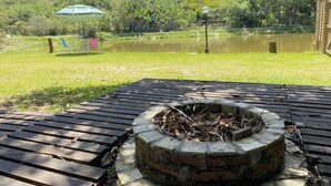 A wooden deck with a brick fire pit, a pond, and a green umbrella in the background. - Recanto Elter (Piracaia)