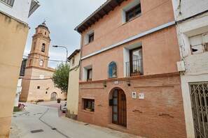 A narrow street with a historic building featuring a bell tower, a balcony with a statue, and a sign indicating a route.