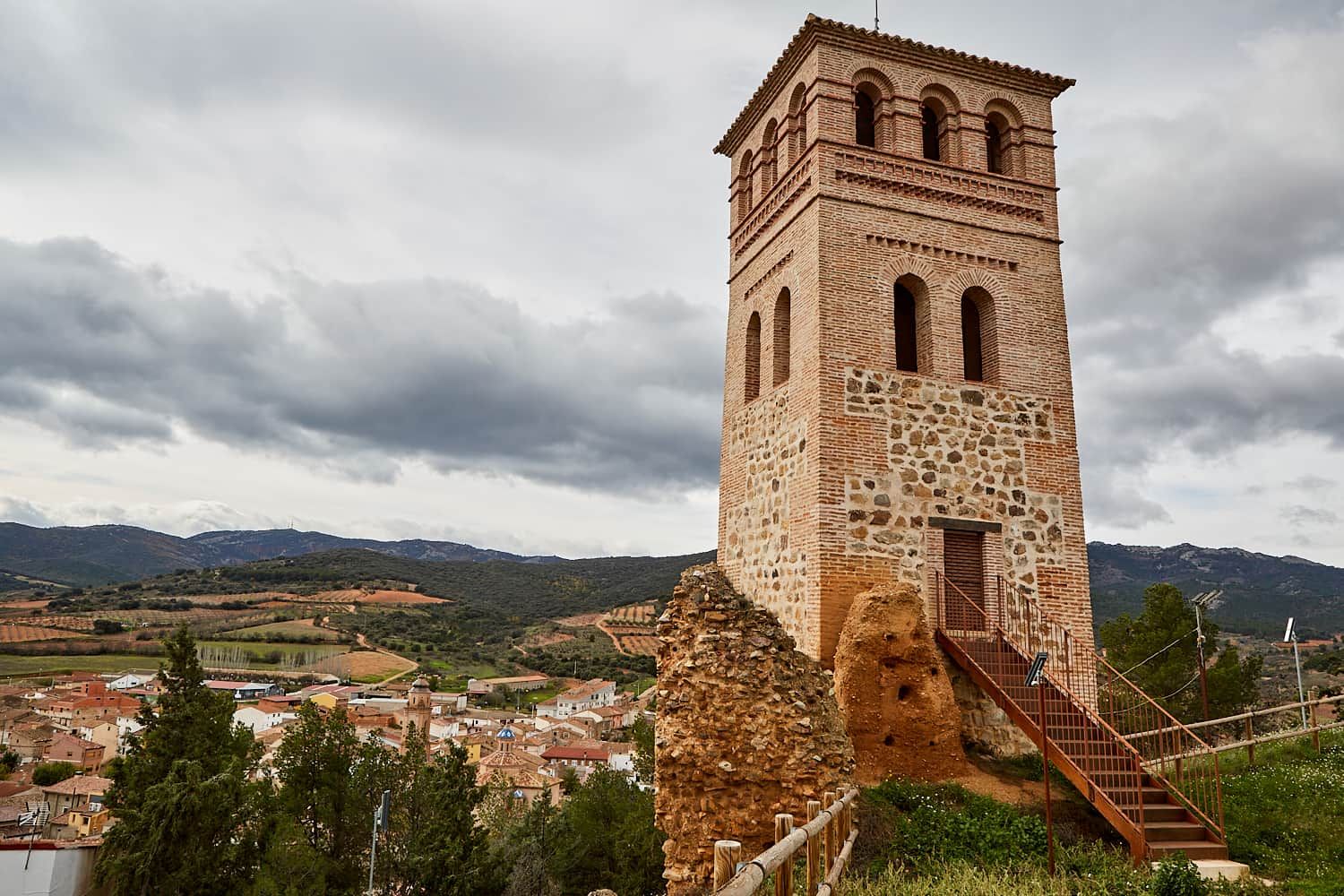 A historic tower with a staircase leading to a door, surrounded by a rural landscape with fields and a village.