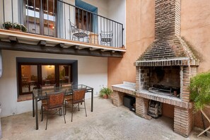 An outdoor patio with a glass table and chairs, a brick barbecue grill, and a balcony with chairs.