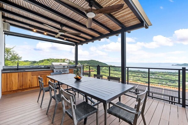 An outdoor deck with a table and chairs, a grill, and a view of a green landscape.