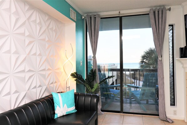 A living room with a black leather sofa, a teal pillow, a geometric wall design, a large sliding glass door, and a view of outdoor furniture.