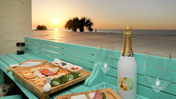 Una mesa con una variedad de quesos, embutidos y galletas, acompañada de una botella de champán y dos copas, con vista a una playa al atardecer.