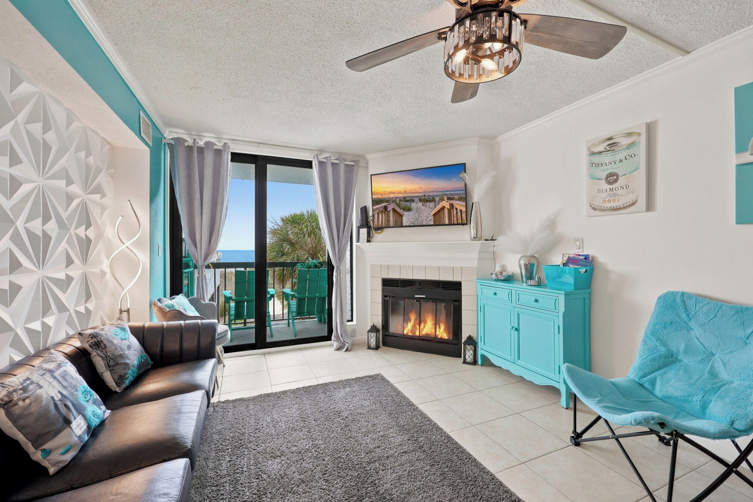 A living room with a fireplace, a sofa, a teal armchair, and a view of the beach through the glass doors.