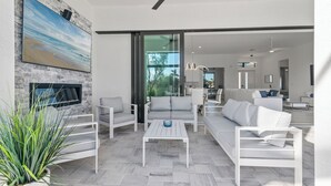 A modern living room with a fireplace, white furniture, and a large painting of a beach scene. - Intervillas Florida - Villa Penelope (Cape Coral)