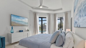 A modern bedroom with a large bed, a ceiling fan, a view of the outdoors, and a painting on the wall. - Intervillas Florida - Villa Penelope (Cape Coral)