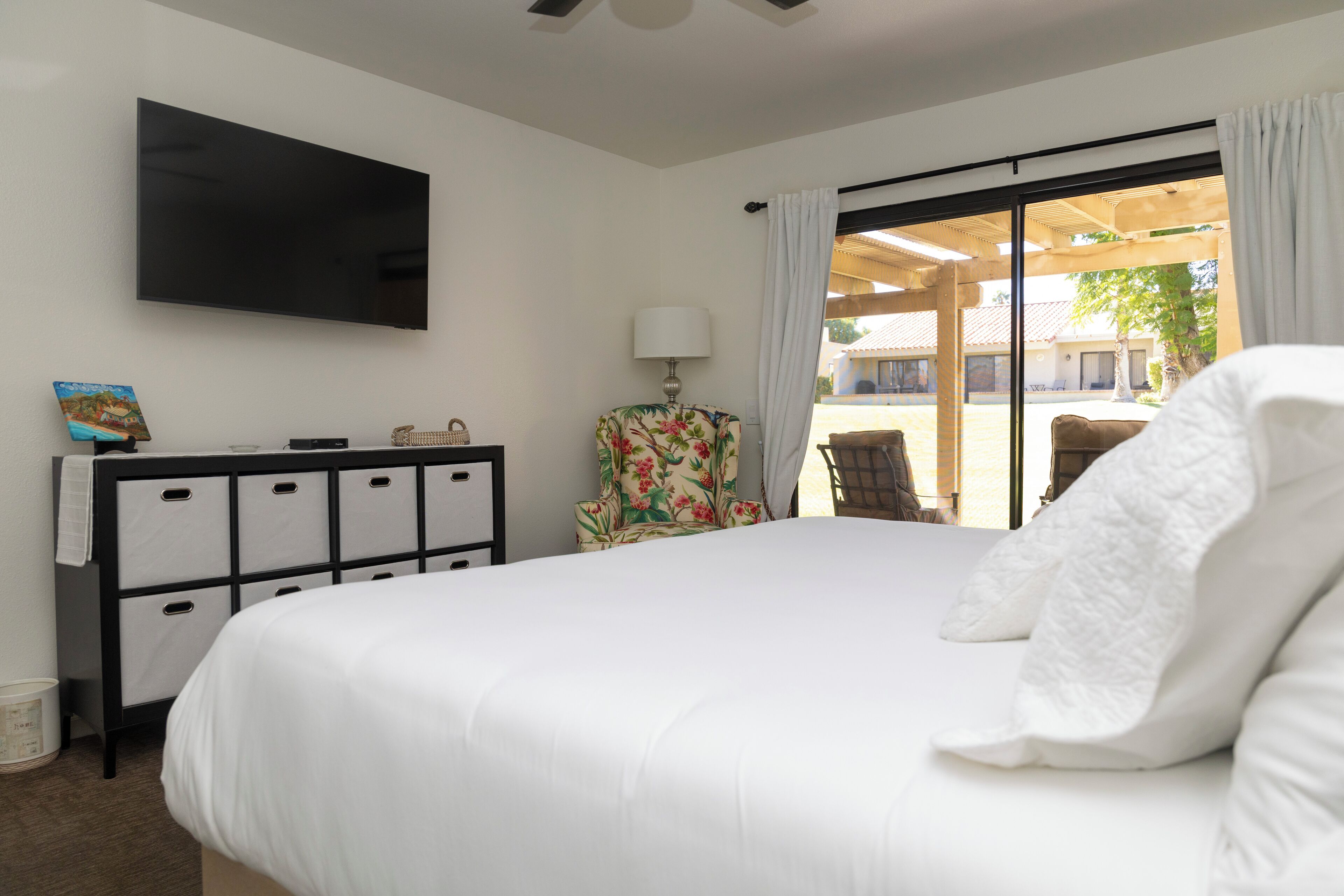A hotel room with a large bed, a flat-screen TV mounted on the wall, a desk with drawers, a chair with a floral pattern, and a view of the outdoor patio through a sliding glass door.