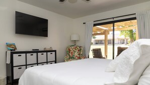 A hotel room with a large bed, a flat-screen TV mounted on the wall, a desk with drawers, a chair with a floral pattern, and a view of the outdoor patio through a sliding glass door. - Charming 2-bedroom condo with AC and WiFi in delightful Palm Desert (Palm Desert)