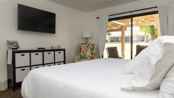 A hotel room with a large bed, a flat-screen TV mounted on the wall, a desk with drawers, a chair with a floral pattern, and a view of the outdoor patio through a sliding glass door.