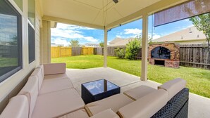 Eine überdachte Terrasse mit einer Couch und einem Tisch, einem Deckenventilator und Blick auf einen Hinterhof mit Kamin aus Ziegelsteinen.