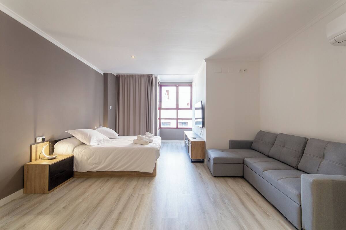 A modern hotel room with a bed, a sofa, a nightstand, and a window with curtains.