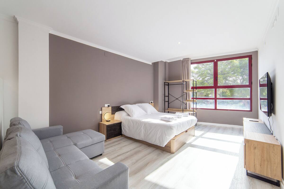 A modern bedroom with a bed, a grey sofa, a wooden nightstand, and a TV.