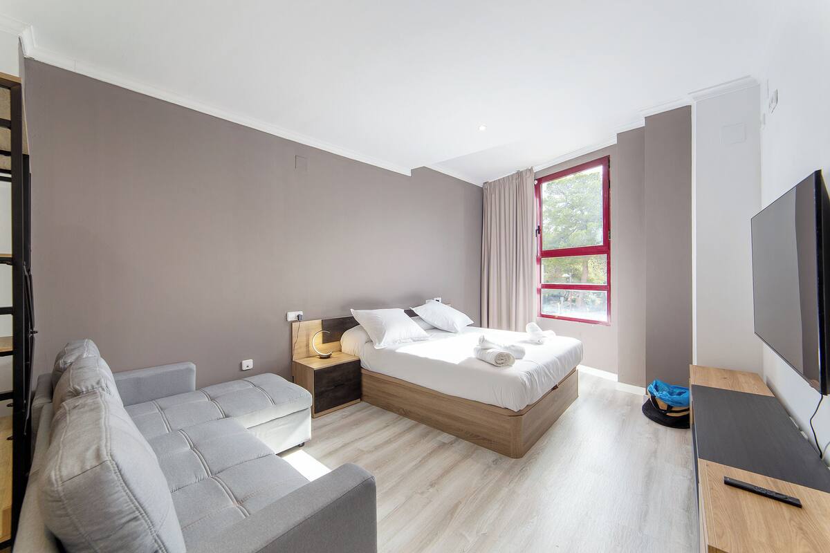 A modern bedroom with a bed, a sofa, a TV, and a window with curtains.