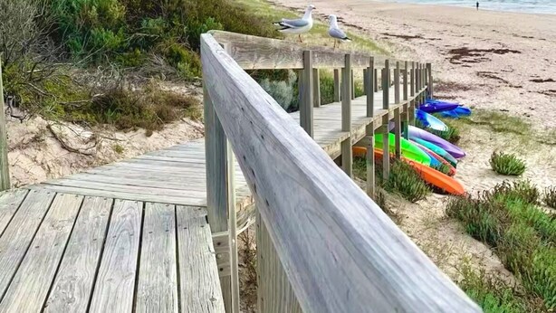 Ein hölzerner Steg führt zu einem Strand, wo bunte Kajaks aneinandergereiht sind.