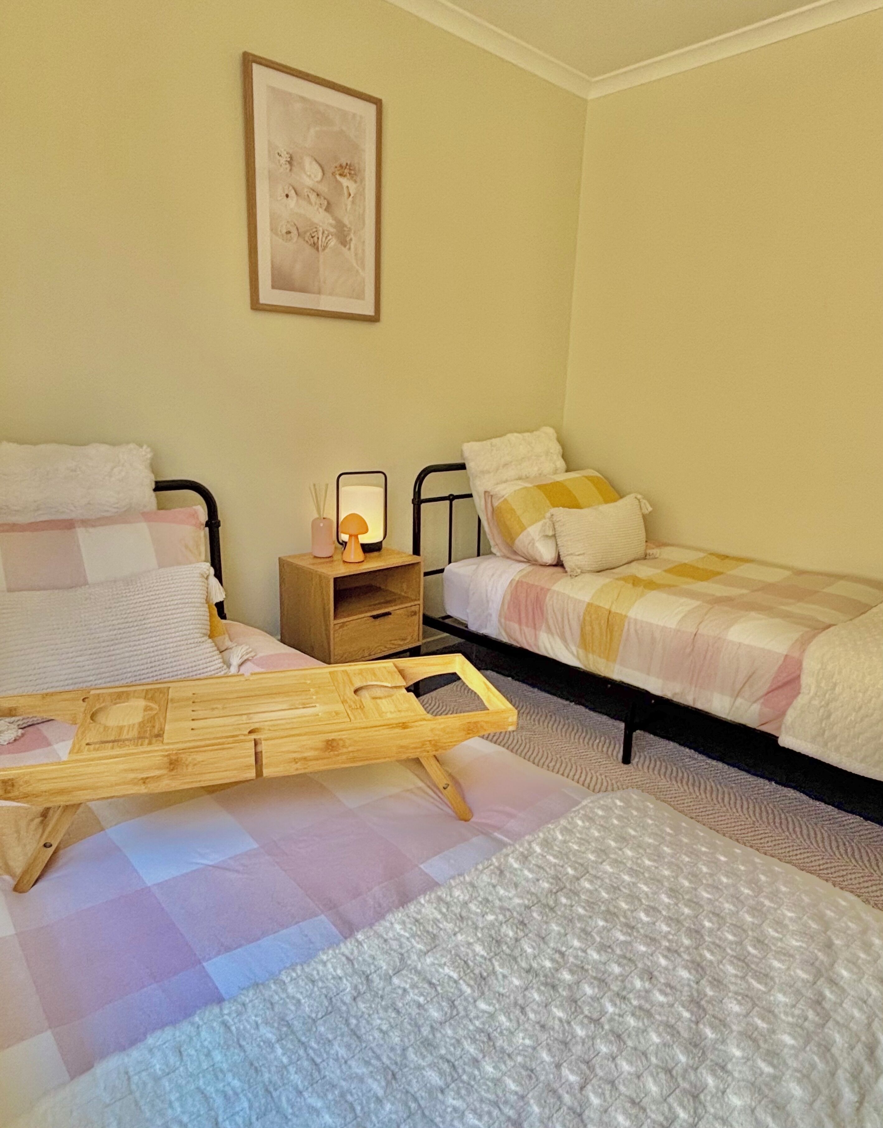 Ein Schlafzimmer mit zwei Betten, einem hölzernen Nachttisch mit Lampe, einem Bild an der Wand und einer kleinen Holzablage auf dem Boden.