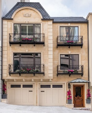 Un immeuble de deux étages avec une façade beige, un toit noir et des balcons en fer forgé décoratifs agrémentés de jardinières.