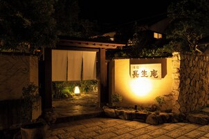 Una entrada de paredes de piedra con un cartel iluminado, una pérgola de madera y un sendero de piedra que lleva a un edificio.