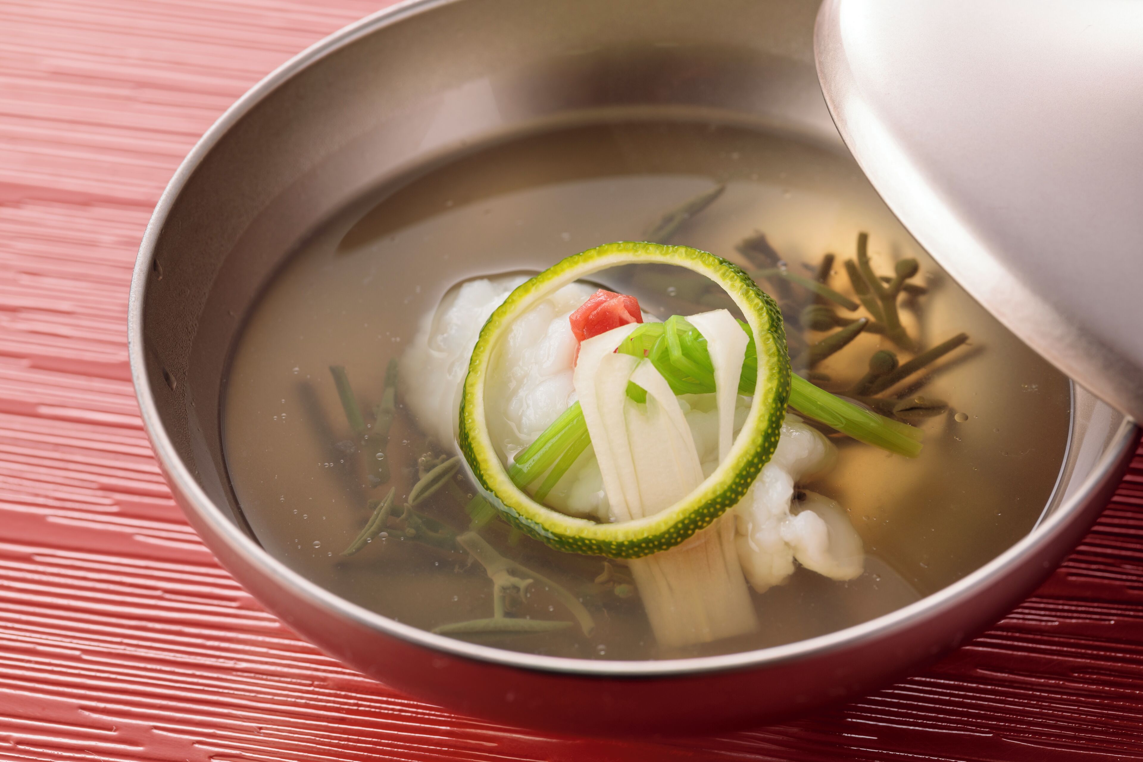 Uma tigela de sopa transparente com vegetais e uma fatia de pepino sobre uma superfície vermelha.