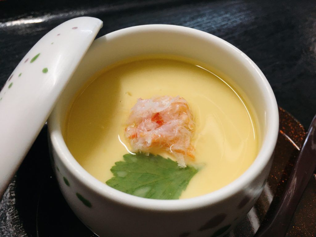 A bowl of soup with a piece of seafood and a garnish, placed on a dark surface.