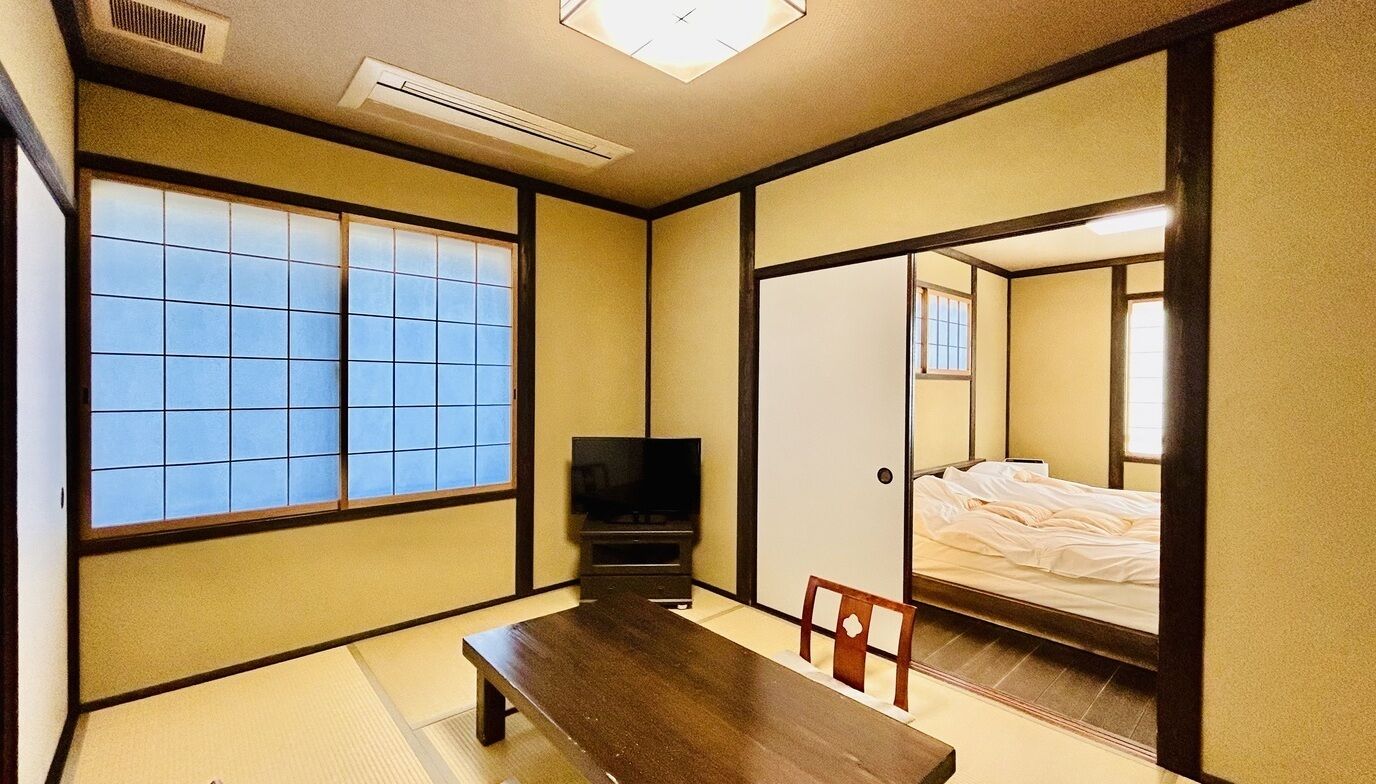 Ein traditioneller japanischer Raum mit Tatami-Böden, einem niedrigen Tisch, einem Fernseher und einem Bett.
