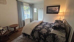 Ein Schlafzimmer mit Bett, Stuhl, kleinem Tisch, Fenster mit Vorhängen und einem Wandbild.