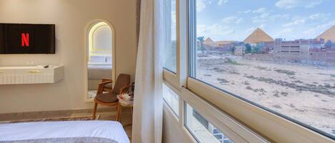 A hotel room with a bed, a TV mounted on the wall, a desk with a chair, and a view of pyramids through the window.
