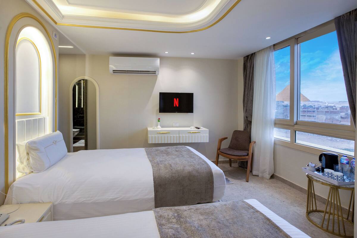 Ein Hotelzimmer mit zwei Betten, einem Fernseher, einem Schreibtisch, einem Sessel und Blick auf die Stadt.