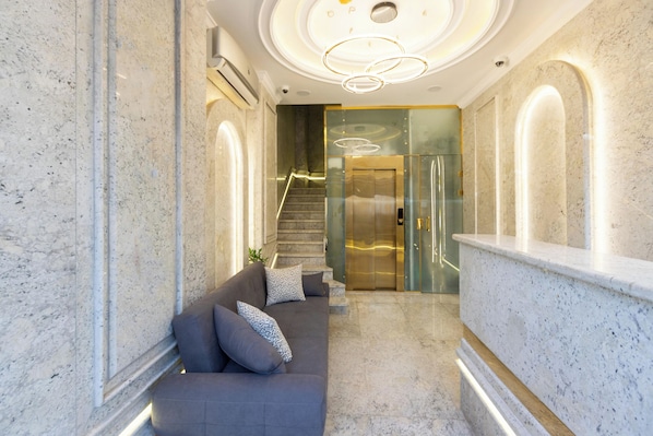 A modern lobby with a marble reception desk, a grey sofa with patterned cushions, and a staircase leading to an elevator. - Pyramids Plateau Hotel (Giza)