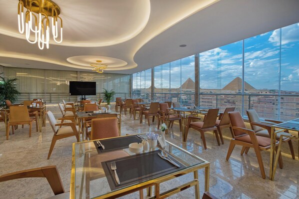 A modern hotel lobby with a large TV, comfortable seating, and a view of the pyramids. - Pyramids Plateau Hotel (Giza)
