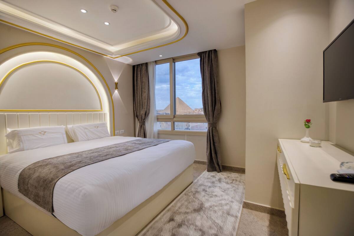 Ein Hotelzimmer mit einem großen Bett, einem Schreibtisch, einem Fernseher und Blick auf eine Pyramide.