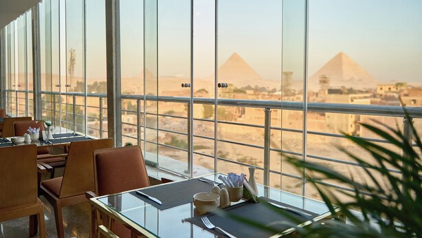A dining area with tables and chairs, large windows offering a view of the pyramids. - Pyramids Plateau Hotel (Giza)