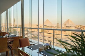A dining area with tables and chairs, large windows offering a view of the pyramids. - Pyramids Plateau Hotel (Giza)