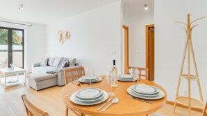Un espace repas moderne avec une table ronde en bois, dressée pour quatre personnes, des murs blancs, un canapé gris et un porte-manteau en bois.