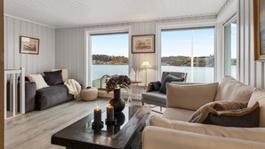 A cozy living room with a view of a lake, featuring a sofa, armchairs, a coffee table with candles, and framed artwork on the walls.
