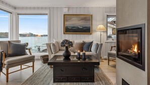 A cozy living room with a fireplace, a large window overlooking a body of water, and a painting on the wall.