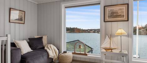 A cozy room with a grey sofa, a small round table with a lamp, and a view of a lake through large windows.